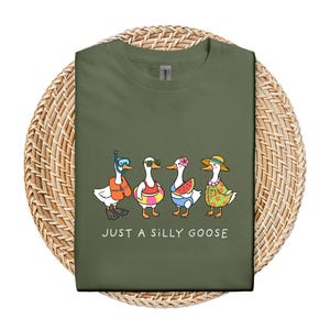 May include: Olive green t-shirt featuring a cartoon illustration of four geese dressed for summer. One goose wears a snorkel, another sunglasses and a life preserver, a third holds a watermelon slice, and the last wears a straw hat. The text "JUST A SILLY GOOSE" is below.