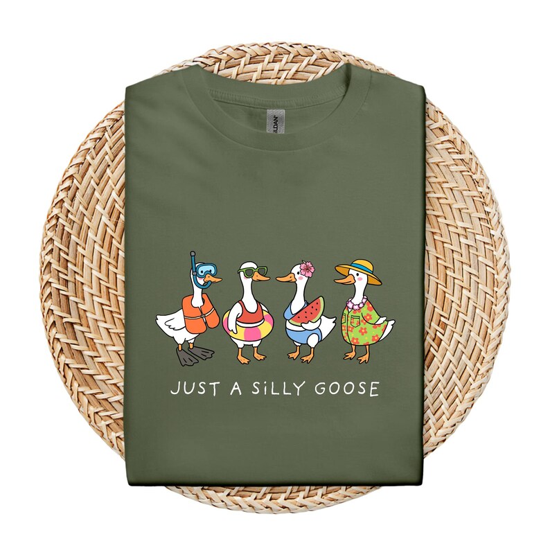 May include: Olive green t-shirt featuring a cartoon illustration of four geese dressed for summer. One goose wears a snorkel, another sunglasses and a life preserver, a third holds a watermelon slice, and the last wears a straw hat. The text "JUST A SILLY GOOSE" is below.