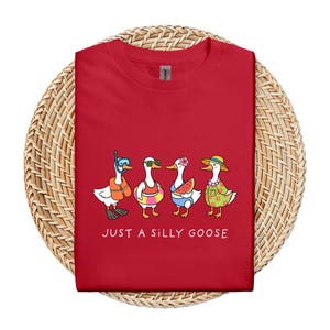 May include: Red t-shirt featuring a whimsical illustration of four cartoon geese in summer attire. One goose wears a snorkel, another sunglasses, a third holds watermelon, and the last wears a floral shirt and hat. The text "JUST A SILLY GOOSE" is printed below.