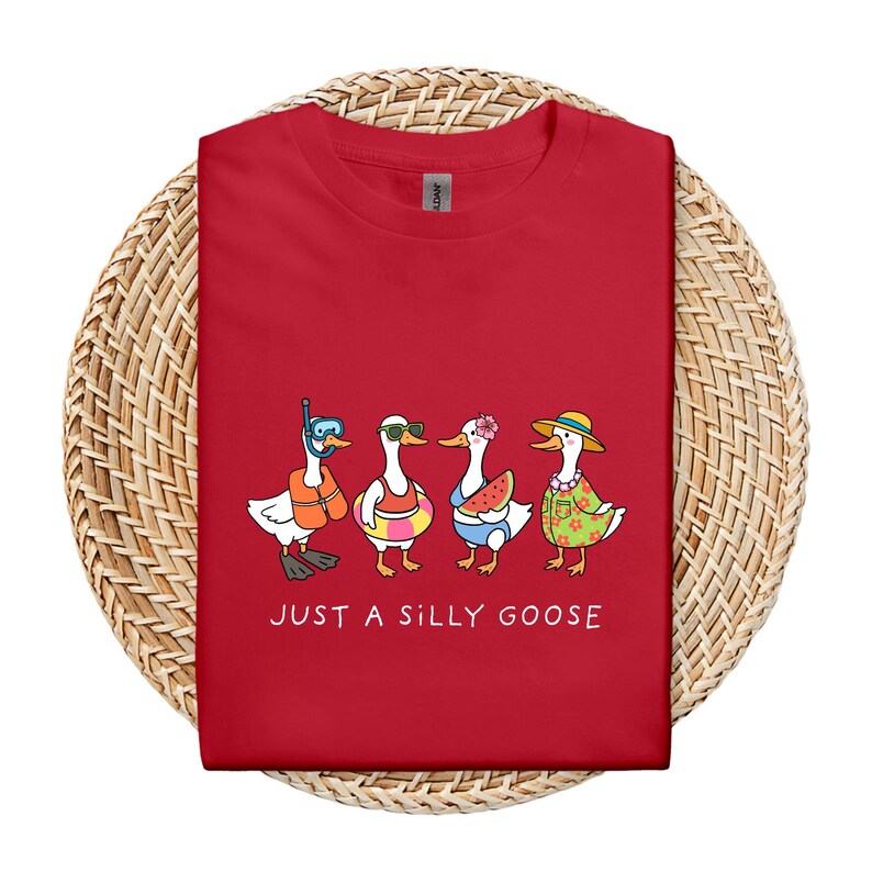 May include: Red t-shirt featuring a whimsical illustration of four cartoon geese in summer attire. One goose wears a snorkel, another sunglasses, a third holds watermelon, and the last wears a floral shirt and hat. The text "JUST A SILLY GOOSE" is printed below.