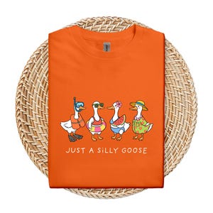 May include: Orange t-shirt featuring a whimsical illustration of four cartoon geese in summer attire. One goose wears a snorkel, another sunglasses, a third a flower, and the last a sun hat. The text "JUST A SILLY GOOSE" is printed below the geese.