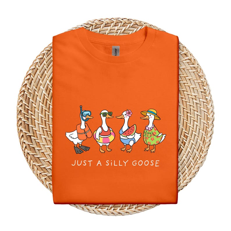 May include: Orange t-shirt featuring a whimsical illustration of four cartoon geese in summer attire. One goose wears a snorkel, another sunglasses, a third a flower, and the last a sun hat. The text "JUST A SILLY GOOSE" is printed below the geese.