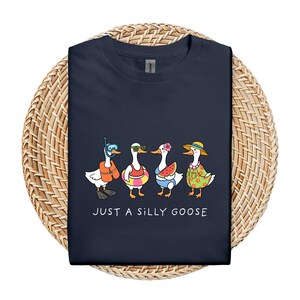 May include: Navy blue t-shirt featuring a whimsical illustration of four geese in summer attire. One goose wears a snorkel, another sunglasses, a third holds a watermelon slice, and the last wears a Hawaiian shirt and hat. The text "JUST A SILLY GOOSE" is printed below.