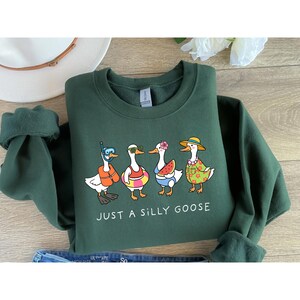 May include: A dark green sweatshirt with the phrase "JUST A SILLY GOOSE" and a graphic of four cartoon geese in summer attire. One goose wears a snorkel and life vest, another has sunglasses and a floatie, the third holds a watermelon slice, and the last wears a hat and floral shirt.