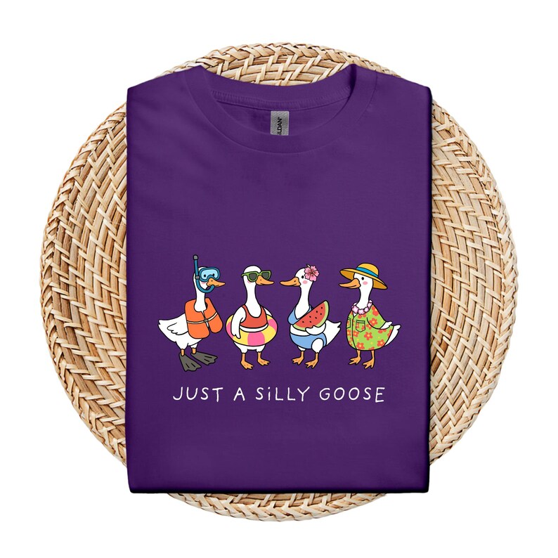 May include: A purple t-shirt featuring a graphic of four cartoon geese, each accessorized with summer items like sunglasses, a life preserver, and a floral lei. The text "JUST A SILLY GOOSE" is printed below the geese.