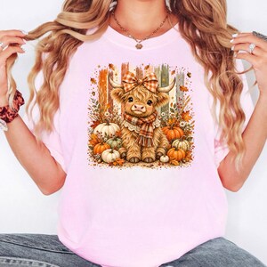 May include: A light pink t-shirt featuring a graphic print of a fluffy Highland cow wearing a plaid bow and scarf. The cow is surrounded by autumn elements including orange and white pumpkins, fall leaves, and vertical wooden-style stripes.