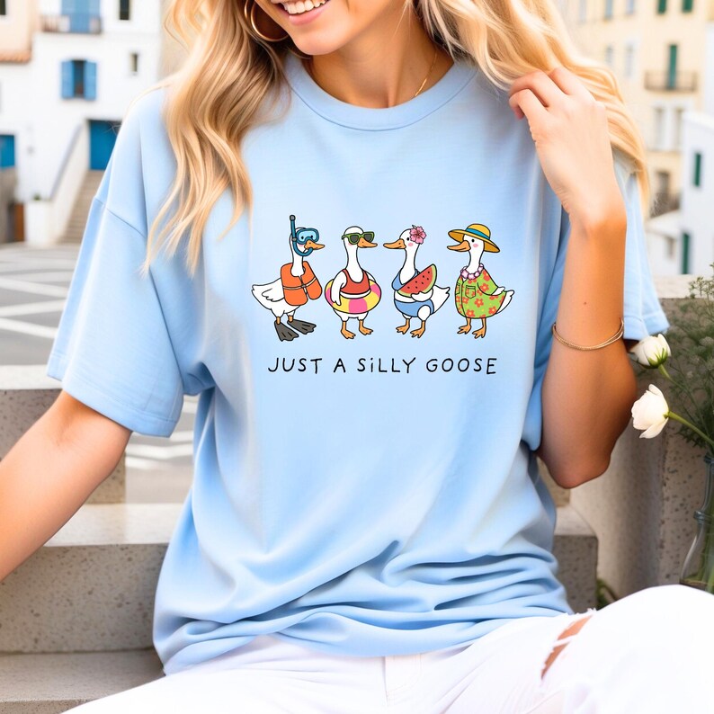 May include: Light blue t-shirt featuring a whimsical illustration of four cartoon geese, each adorned with summer accessories like sunglasses, life vests, and hats. The text "JUST A SILLY GOOSE" is printed below the geese. The shirt is made of a soft material.