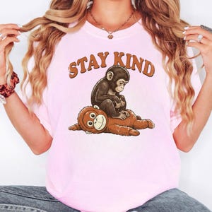 May include: A light pink t-shirt featuring a graphic of a monkey sitting on top of a stuffed animal monkey. The text "STAY KIND" is printed above the monkeys in brown letters. The shirt is made of a soft material.