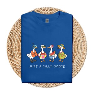 May include: Royal blue t-shirt featuring a whimsical illustration of four cartoon geese in summer attire. One goose wears a snorkel, another sunglasses, a third holds a watermelon slice, and the last wears a floral shirt and straw hat. The text "JUST A SILLY GOOSE" is printed below.