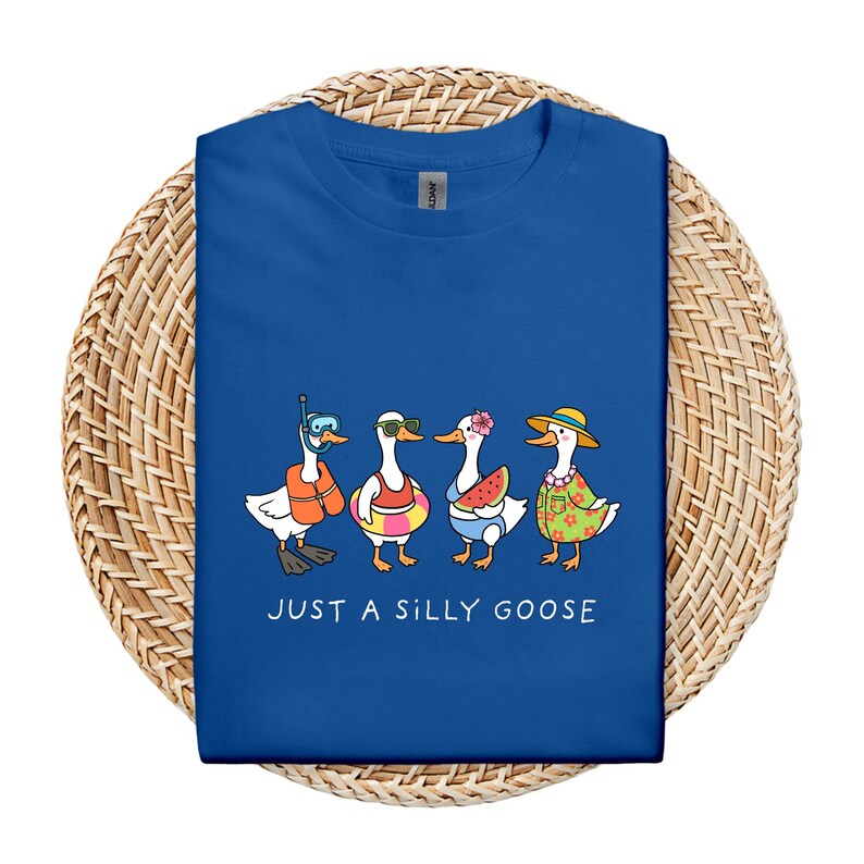 May include: Royal blue t-shirt featuring a whimsical illustration of four cartoon geese in summer attire. One goose wears a snorkel, another sunglasses, a third holds a watermelon slice, and the last wears a floral shirt and straw hat. The text "JUST A SILLY GOOSE" is printed below.