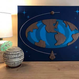 Pode incluir: Uma pintura em tela azul com um recorte de madeira da Terra em órbita ao redor de um pequeno planeta. A pintura também apresenta uma nave espacial e estrelas.