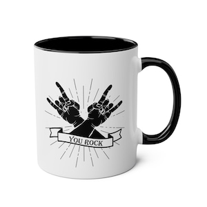 May include: White ceramic mug with a black interior and handle. The mug features a black and white graphic of two hands making the rock and roll hand gesture, with the words "YOU ROCK" on a banner.