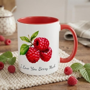 Peut inclure: Un mug en céramique blanc avec un intérieur et une anse rouges, orné d'un motif de trois framboises rouges avec des feuilles vertes et du texte noir "I Love You Berry Much" imprimé sur le devant, centré sous l'illustration des fruits.