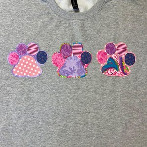 May include: A heather gray t-shirt featuring three appliqued paw prints across the chest. Each paw print is constructed from various fabric patches in shades of pink, purple, and blue, showcasing diverse patterns including polka dots, stripes, and floral motifs.