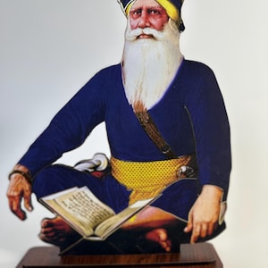 May include: A wooden cutout of a man in a blue turban and robe, seated cross-legged, reading a book. The figure has a long white beard and a yellow sash. The cutout is mounted on a brown wooden base.