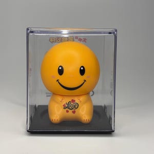 May include: Orange smiley face figurine in a clear plastic display case. The figurine has black eyes and a black smile. The word "Love" is written on the chest of the figurine. The figurine is on a black base.