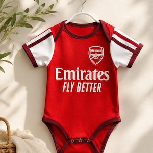 May include: A red and white baby bodysuit featuring the Arsenal football club crest on the chest and the text Emirates FLY BETTER in white lettering. The garment has short sleeves with dark red trim and a snap-button closure at the bottom.