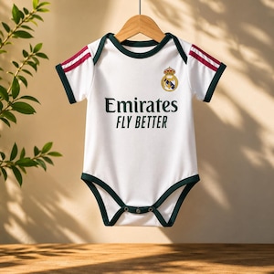 May include: White baby bodysuit with green trim and red stripes on the sleeves. The bodysuit features the text "Emirates FLY BETTER" and a team crest. The garment is hanging on a wooden hanger.