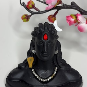 May include: Black Adiyogi statue with a red forehead mark, gold snake, and jewelled necklace. The statue has long hair and a white crescent moon. Pink flowers and branches are in the background. The word 'Adiyogi' is written on the base.