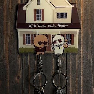 May include: A house-shaped key holder with the text "Rich Dudu Bubu House". The key holder features two cartoon bears wearing sunglasses. Two sets of keys hang from the key holder, which is mounted on a dark wooden surface.