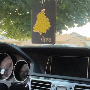 Puede incluir: Un ambientador rectangular para coche con un mapa dorado de Punjab sobre un fondo negro, colgado de un hilo plateado. La palabra "Punjab" está escrita en oro debajo del mapa. El ambientador cuelga frente al salpicadero de un coche.
