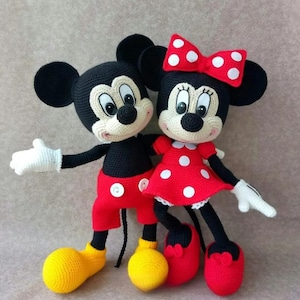 May include: Crocheted Mickey Mouse and Minnie Mouse dolls. Mickey wears red shorts, yellow shoes, and white gloves. Minnie wears a red dress with white polka dots, a red bow, and red shoes. Both dolls have black ears and smiling faces.