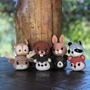 May include: A collection of handmade crochet animal toys, including an owl, beaver, rabbit, bear, raccoon, and fox. Each toy features detailed facial features and is made with various yarn colors. The toys are arranged on a wooden surface.
