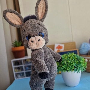 Puede incluir: Un juguete de peluche de burro gris hecho a mano con orejas grandes y ojos azules. El burro tiene un hocico beige claro y pezuñas negras. El juguete está sobre una superficie azul. Es un peluche lindo y tierno.