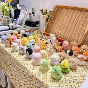 May include: A collection of small, colorful, crocheted animal figurines, including bears, pandas, and other creatures. The handmade toys are displayed on a patterned tablecloth, with a woven basket in the background.