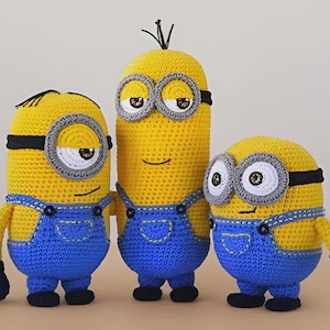 Könnte beinhalten: Drei handgefertigte Minion-Figuren aus Häkelarbeit. Jeder Minion ist gelb mit blauen Latzhosen, schwarzen Handschuhen und grauen Brillen. Die Figuren variieren in der Höhe, wobei eine größer ist als die anderen. Sie haben schwarze Haare und lächelnde Gesichter.