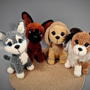 Op de afbeelding: Vier handgemaakte gehaakte hondenknuffels in verschillende kleuren, waaronder grijs, bruin en beige. Elke pluchen hond heeft unieke kenmerken, zoals flaporen en verschillende vachtpatronen. Ze worden weergegeven op een rond, geweven oppervlak.