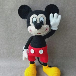 Könnte beinhalten: Handgemachte Mickey Mouse-Puppe aus Häkelarbeit. Die Puppe hat einen schwarzen Kopf mit großen runden Ohren, einem weißen Gesicht und einer schwarzen Nase. Sie trägt einen schwarzen Körper, rote Shorts mit weißen Knöpfen, weiße Handschuhe und gelbe Schuhe. Die Puppe winkt.