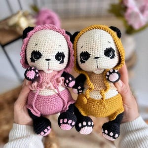 Op de afbeelding: Twee handgemaakte gehaakte panda's. Eén panda draagt een roze outfit met capuchon en de andere een gele outfit met capuchon. Beide panda's hebben zwart-witte gezichten, zwarte poten met roze kussentjes en zijn ongeveer 15 cm hoog.