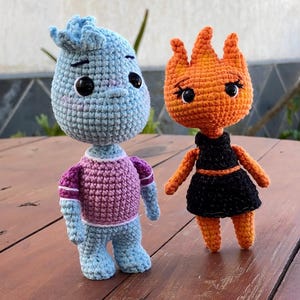 Op de afbeelding: Twee handgemaakte gehaakte poppen. Eén is blauw met een paarse en roze trui, de andere is oranje met een zwarte jurk. Beide poppen hebben grote zwarte ogen en staan op een houten oppervlak.