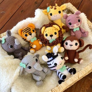 May include: A collection of handmade crochet animal toys, including a giraffe, lion, tiger, zebra, elephant, rhino, monkey, and hippo. Each toy features detailed stitching, soft colors, and small bows. The toys are arranged in a woven basket.