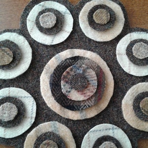 May include: A brown felt flower with multiple layers of circles in shades of brown, beige, and white. The flower has a scalloped edge and is attached to a white ribbon with brown buttons.