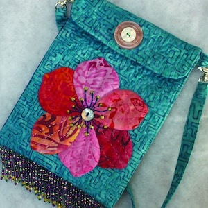 May include: A teal blue fabric crossbody bag with a flap closure and a beaded fringe. The bag features a large pink and red flower applique with a button in the center.