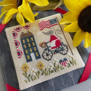 Puede incluir: Patrón de punto de cruz con una bandera estadounidense roja, blanca y azul, una casa pequeña, una bicicleta y flores. El diseño está en una tela de color crema con un fondo gris.