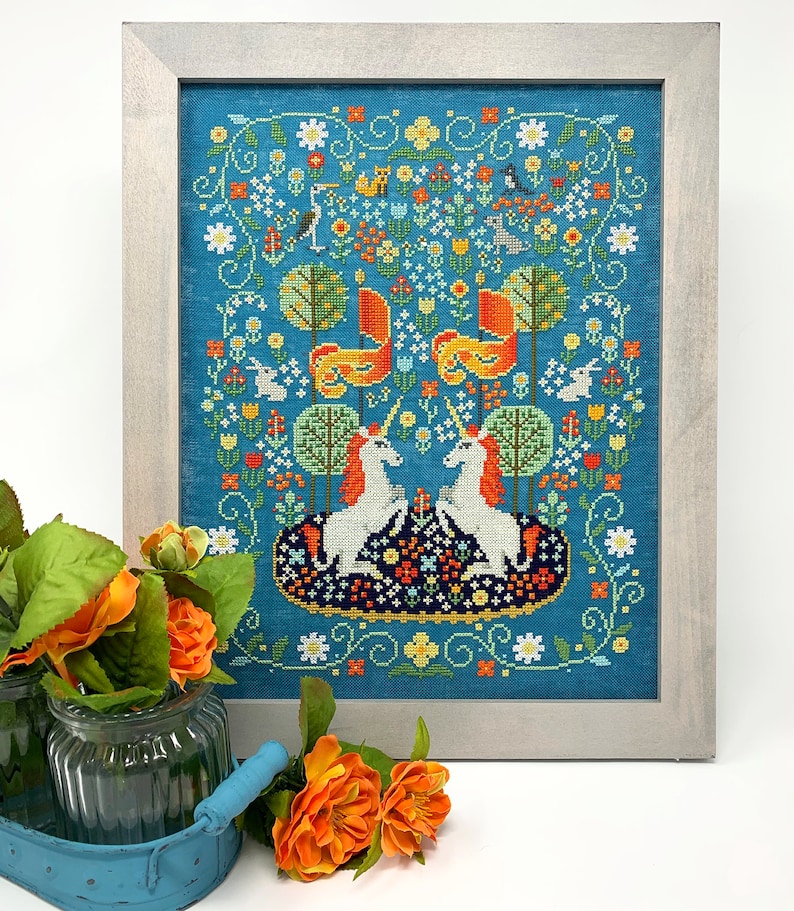 May include: Framed cross-stitch embroidery featuring two white unicorns with orange manes and tails, standing on a blue and orange floral patterned rug. The background is a teal blue with a floral border.