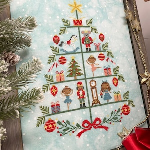 Op de afbeelding: Een kruissteekpatroon van een kerstboom versierd met ornamenten, cadeaus, een notenkraker, een schommelpaard, een ballerina en een klok. De boom is versierd met rode, groene en gouden ornamenten. De achtergrond is lichtblauw met witte sneeuwvlokken.