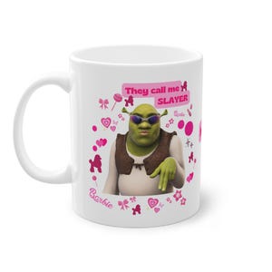 Könnte beinhalten: Weiße Keramiktasse mit einer grünen Ogerfigur, die eine Sonnenbrille und eine braune Weste trägt. Der Text "They call me SLAYER" ist in Pink mit einem pinkfarbenen Barbie-Logo. Der Hintergrund ist weiß mit rosa Herzen, Schleifen und Poodlesilhouetten.