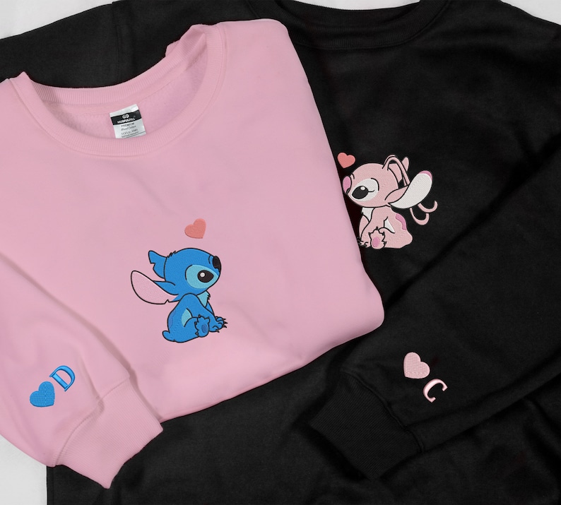 May include: Two pink and black sweatshirts with a blue and pink cartoon character design. The pink sweatshirt has a blue Stitch character with a heart above it. The black sweatshirt has a pink Angel character with a heart above it. Both sweatshirts have a heart with a letter on the sleeve.