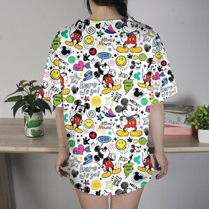 May include: White t-shirt dress with a colorful Mickey Mouse pattern. The design includes Mickey Mouse illustrations, stars, hearts, and text like "Here we go!" and "Mickey Mouse". The dress has short sleeves and a relaxed fit.