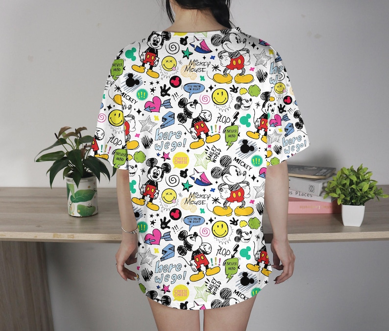 May include: White t-shirt dress with a colorful Mickey Mouse pattern. The design includes Mickey Mouse illustrations, stars, hearts, and text like "Here we go!" and "Mickey Mouse". The dress has short sleeves and a relaxed fit.
