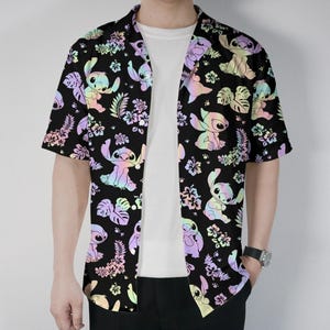 Könnte beinhalten: Schwarzes kurzärmeliges Hemd mit einem farbenfrohen Muster aus Zeichentrickfiguren und tropischen Blumen. Das Hemd mit klassischem Kragen wird offen über einem weißen T-Shirt getragen. Das Design verwendet Pastellfarben.