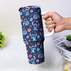 Puede incluir: Un vaso grande con un asa blanca, con un fondo azul marino con un patr&oacute;n repetido del personaje Stitch, flores y hojas. El vaso est&aacute; sostenido por una mano.