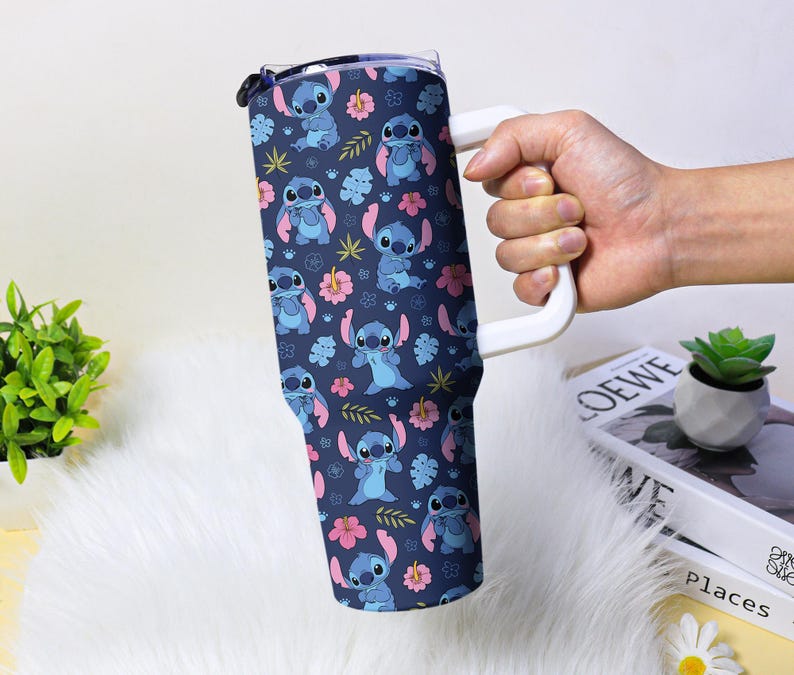 Puede incluir: Un vaso grande con un asa blanca, con un fondo azul marino con un patr&oacute;n repetido del personaje Stitch, flores y hojas. El vaso est&aacute; sostenido por una mano.