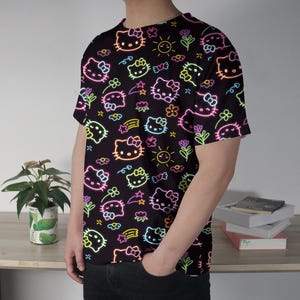 May include: Black t-shirt featuring a neon-style pattern of Hello Kitty characters, flowers, stars, and suns. The design includes pink, yellow, green, and blue outlines on a black background. The short-sleeved shirt is made of a soft material.