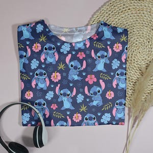 Pu&ograve; includere: Una maglietta blu navy con un motivo ripetuto del personaggio Stitch, fiori rosa e foglie verdi. La maglietta &egrave; piegata e posizionata su una superficie rosa chiaro con cuffie.