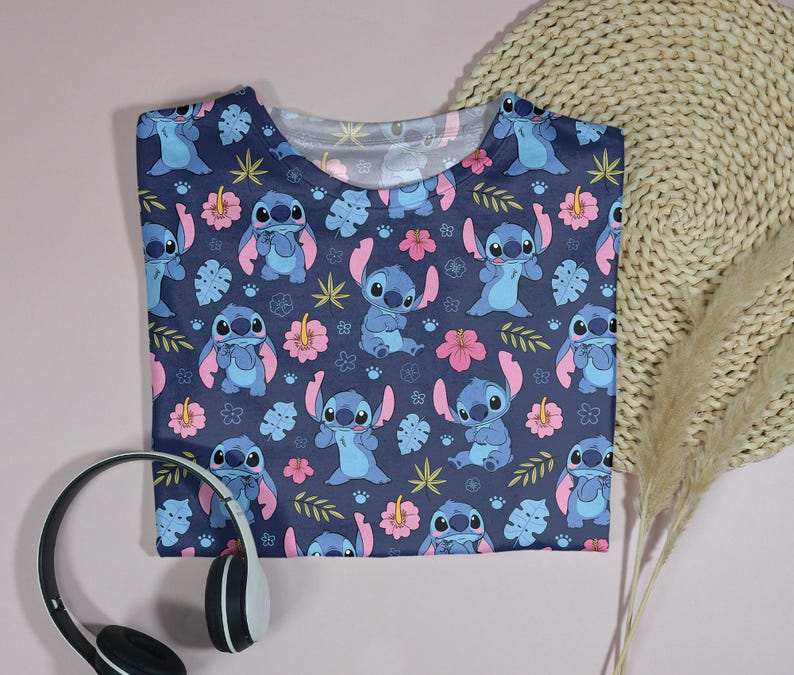Pu&ograve; includere: Una maglietta blu navy con un motivo ripetuto del personaggio Stitch, fiori rosa e foglie verdi. La maglietta &egrave; piegata e posizionata su una superficie rosa chiaro con cuffie.